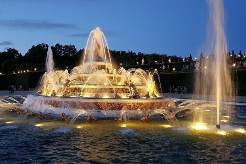 A multi-tiered fountain with water jets and lights, surrounded by a body of water and a landscaped area with trees. 1