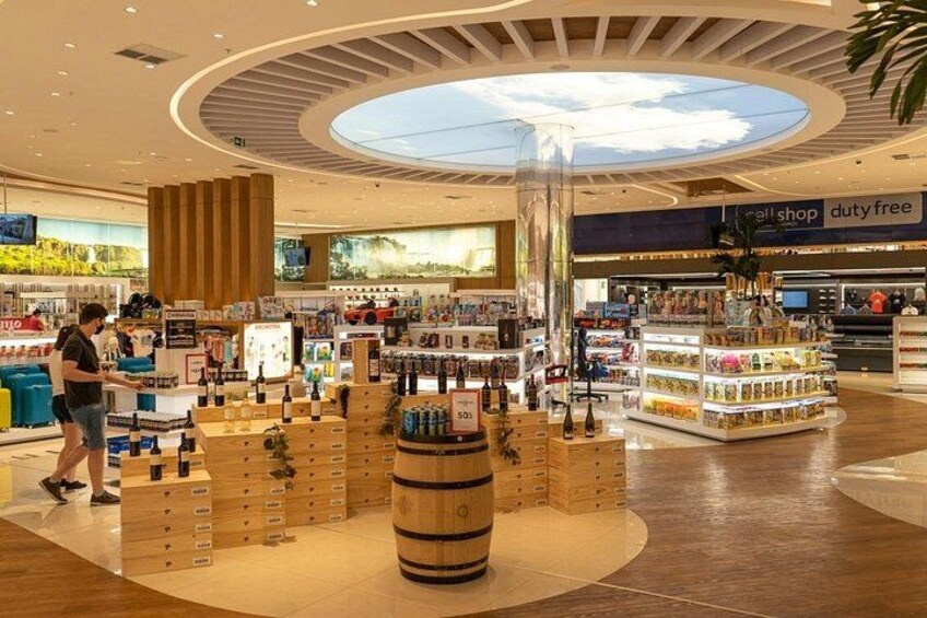 Un centro comercial moderno con una zona de tiendas libres de impuestos, diversas tiendas minoristas y una exhibición central con botellas de vino y un barril. 3