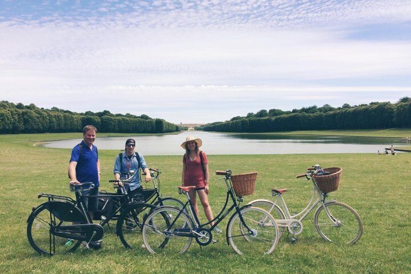 Tre persone in piedi su un prato con biciclette, un lago e alberi sullo sfondo. 3