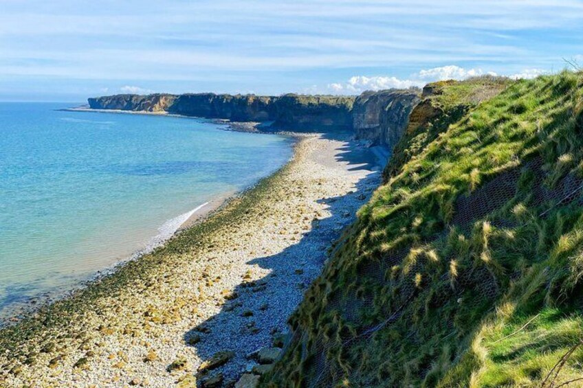 Paesaggio costiero con spiaggia di ciottoli, scogliere erbose e mare azzurro cristallino. 1