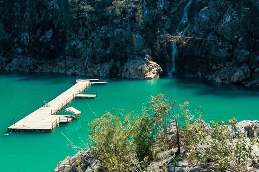 Un lago turquesa con un muelle de madera que se adentra en él, rodeado de acantilados rocosos y una cascada. 2