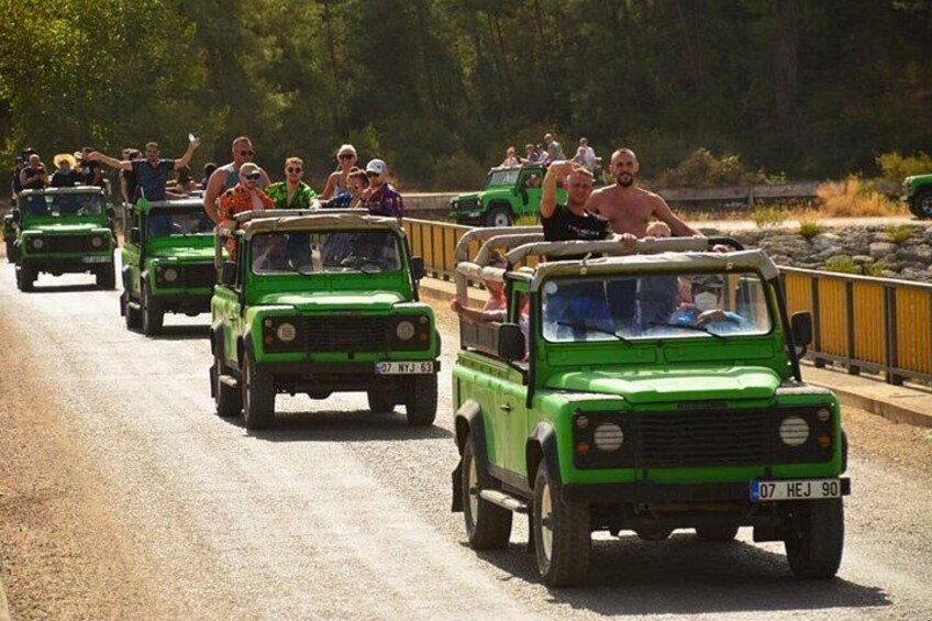 A convoy of green off-road vehicles on a paved road with passengers. 2