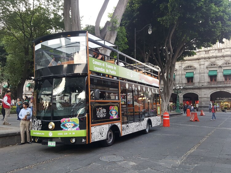A double-decker bus with a colorful mural and text in Spanish, parked on a city street with pedestrians and trees in the background. 1