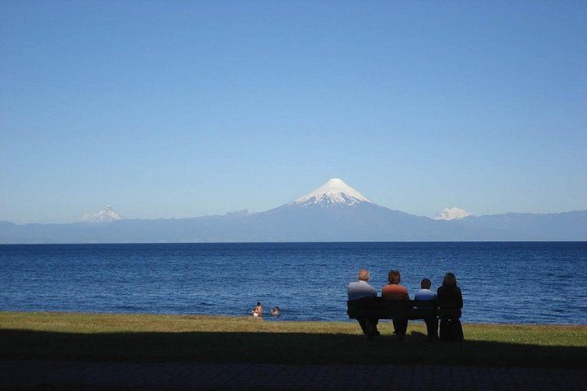 A group of people sitting on a bench by the water, a mountain in the background, and a grassy area in the foreground. 3
