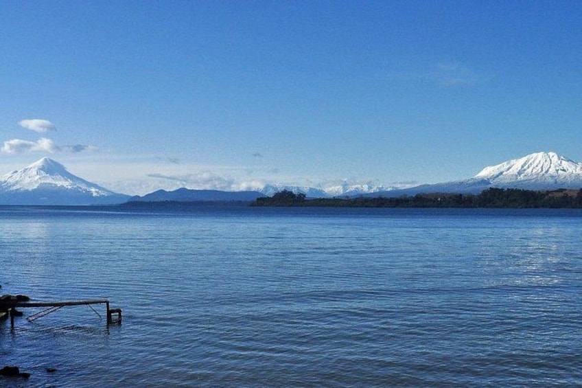 A calm lake with a wooden dock, a snow-capped mountain in the background, and a clear blue sky. 2