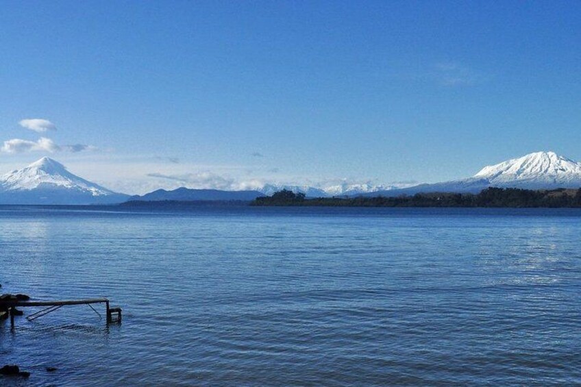 A calm lake with a wooden dock, a snow-capped mountain in the background, and a clear blue sky. 2
