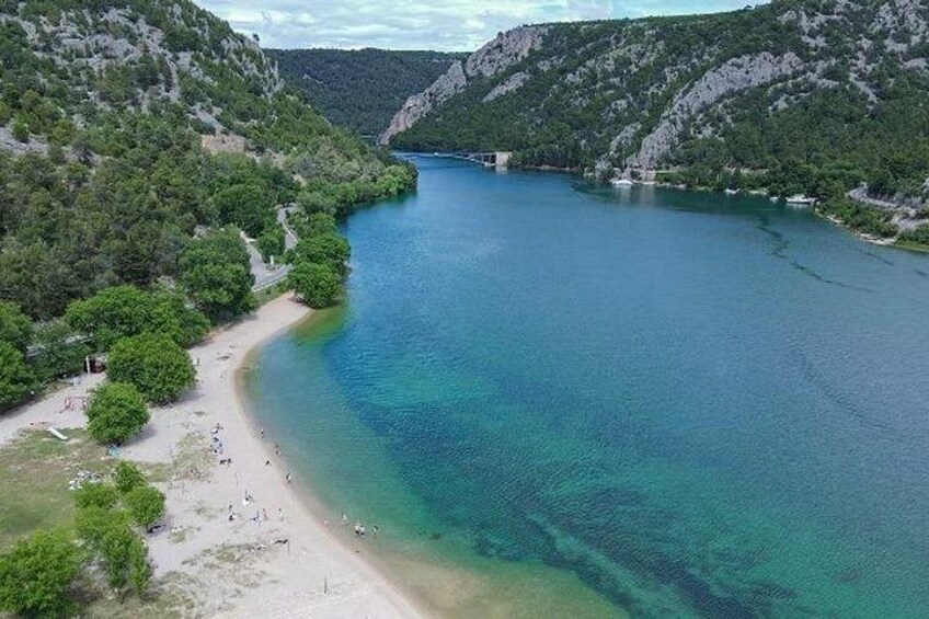 A coastal landscape with a beach, clear blue water, and surrounding hills. 5