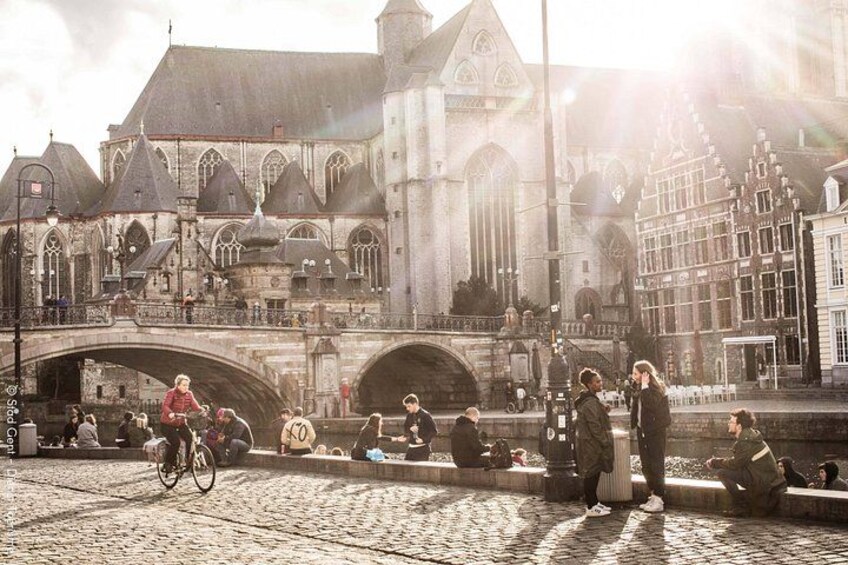 A cobblestone street with a bridge, historic buildings, and people sitting and walking. 2
