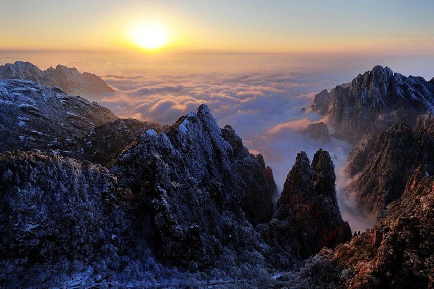 A mountainous landscape with snow-covered peaks, a sun setting or rising in the background, and a layer of clouds below. 5