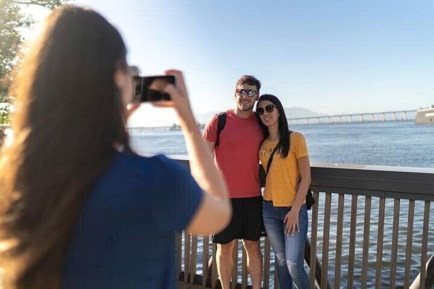 A person taking a photo of a couple standing by a waterfront with a bridge in the background. 3