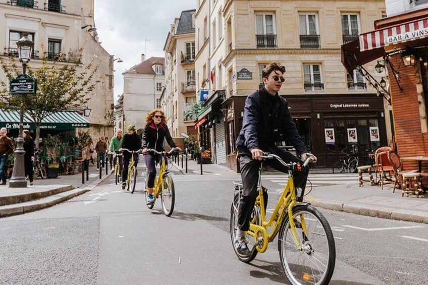 Two people riding bicycles on a city street with buildings and a café in the background. 4