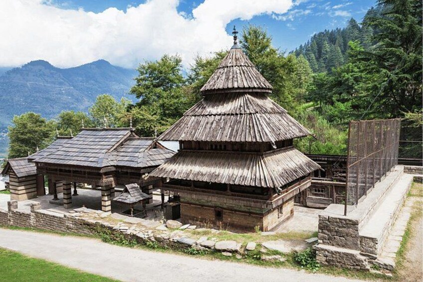 A traditional wooden structure with a conical roof, surrounded by stone steps and a stone wall, set against a mountainous backdrop. 1