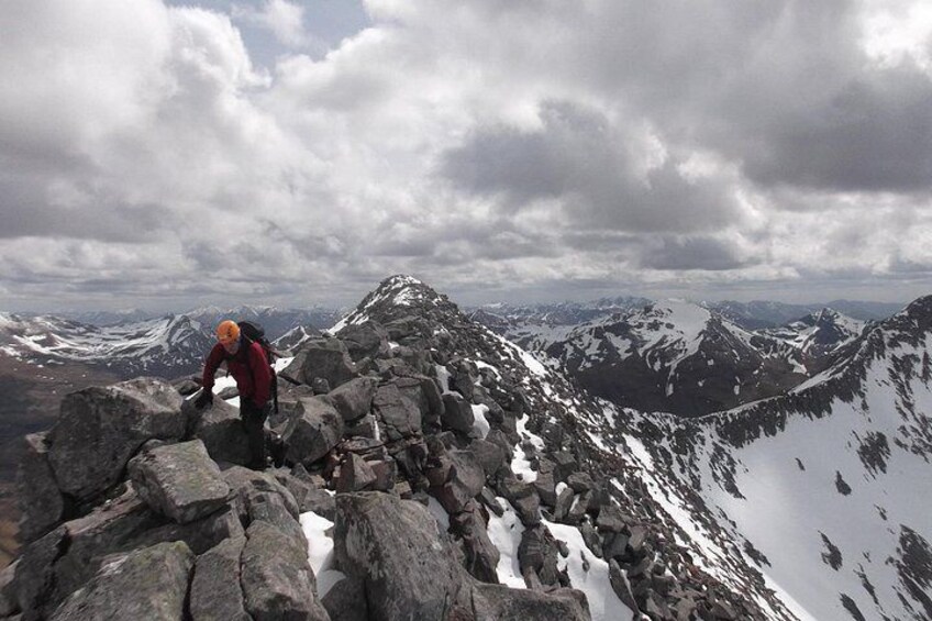 A hiker on a rocky mountain path with snow patches and distant mountain peaks under a cloudy sky. 4