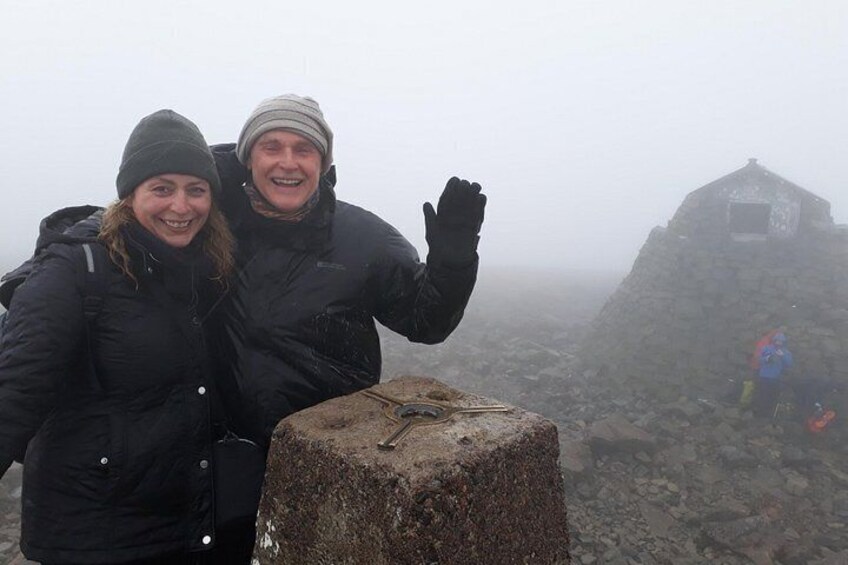 Two people standing on a rocky outcrop with a stone structure in the background. 3