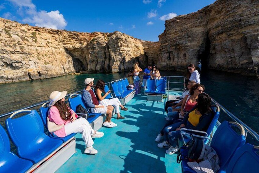 A boat tour with passengers seated on blue chairs, rocky cliffs, and clear blue water. 1