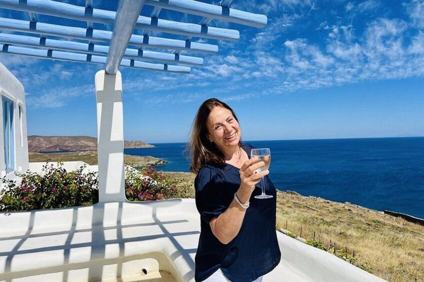 A person holding a glass, standing on a terrace with a view of the sea and a white building with blue accents. 1