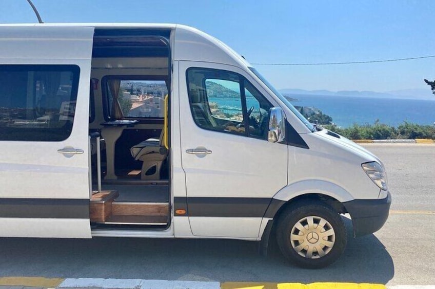 A white van parked on a road with a clear view of the sea and distant mountains in the background. 1