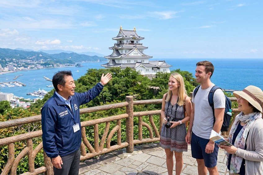 A group of people standing on a wooden observation deck overlooking a coastal city with a prominent castle. 1