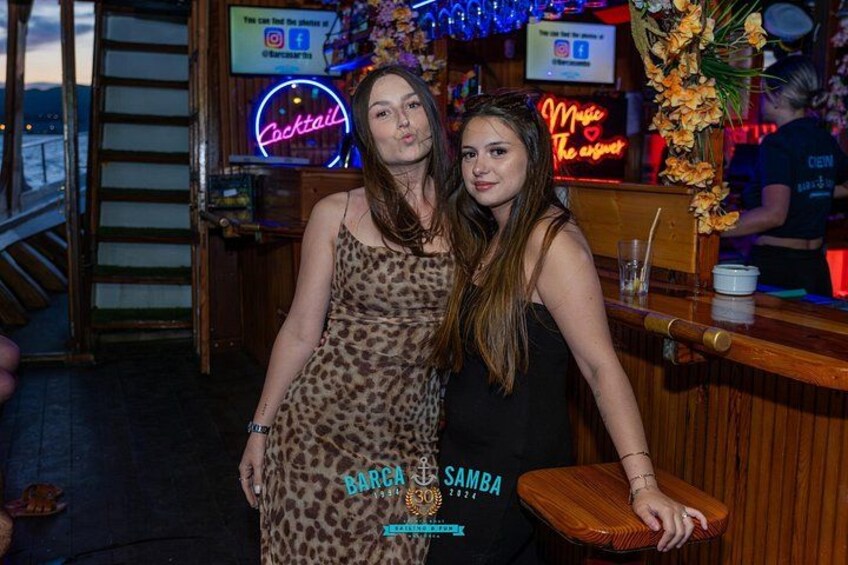 Zwei Personen an einer Bar, mit einem leuchtenden Cocktail-Schild und einem weiteren Schild mit der Aufschrift 'Miss The Essence'. 2