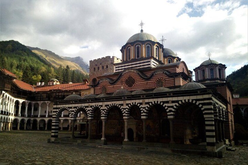 A historic building with a large central dome, arched walkways, and a mountainous backdrop. 1