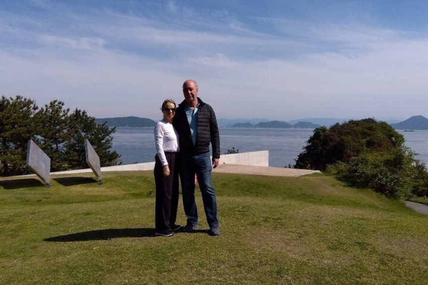Two people standing on a grassy area with a view of the sea and mountains in the background. 1
