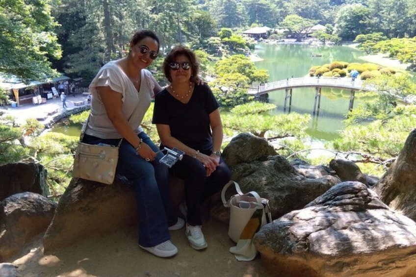 Two people sitting on rocks overlooking a pond with a bridge and Japanese-style landscaping. 1