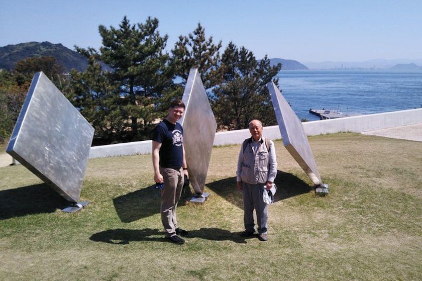 Two people standing in front of large metallic sculptures on a grassy area with a view of the ocean and mountains in the background. 3