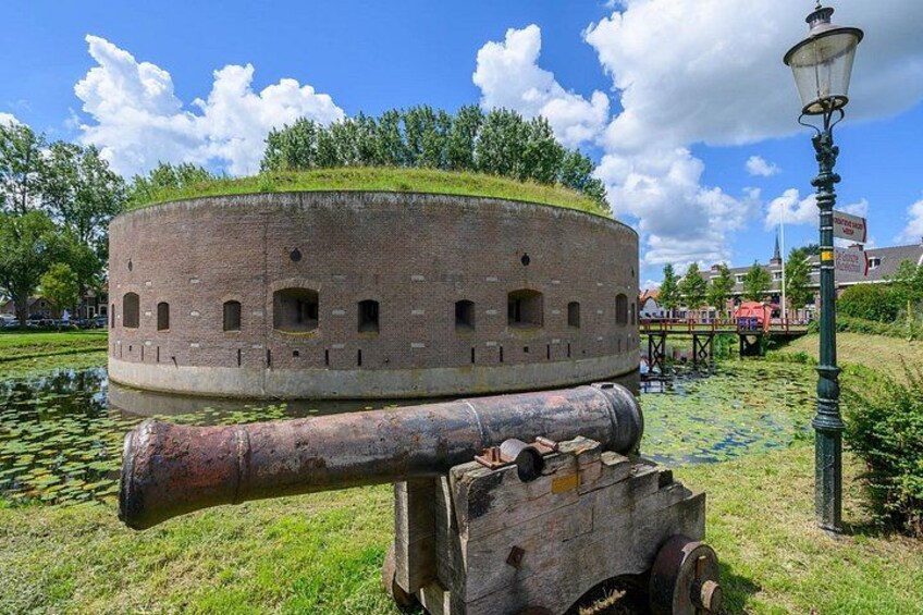 A circular brick structure with a cannon on a wheeled platform, surrounded by a pond with lily pads, under a clear sky. 2