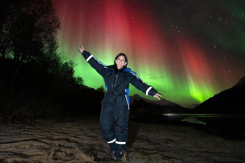 A person standing on a sandy beach with arms outstretched, facing a vibrant aurora display in the night sky. 3