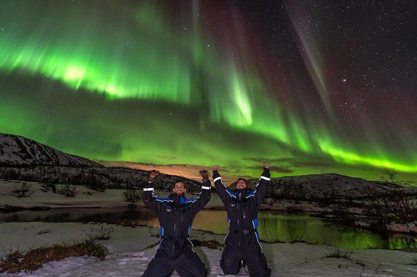 Two people with arms raised in a snowy landscape under the Northern Lights. 1