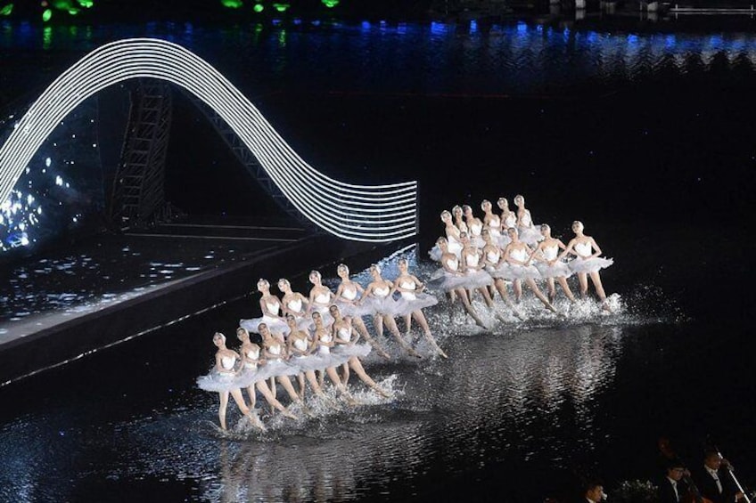 Una función de ballet con bailarines en trajes blancos sobre un escenario con un elemento acuático reflectante y una estructura en forma de ola al fondo. 1