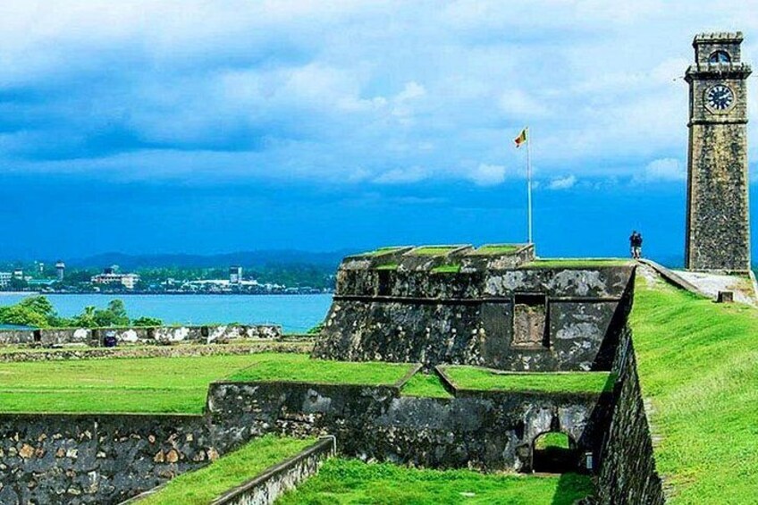 A coastal fort with a clock tower, grassy embankments, and a clear view of the sea. 1
