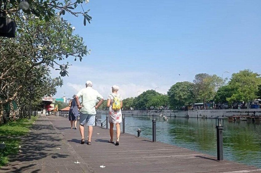 A wooden boardwalk along a river with people walking, trees, and a clear sky. 2