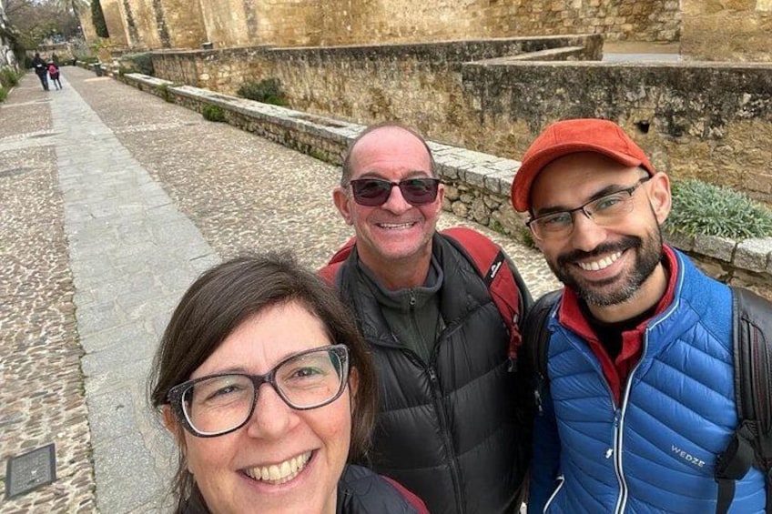 Three people standing on a cobblestone path with stone walls and a stone bridge in the background. 4