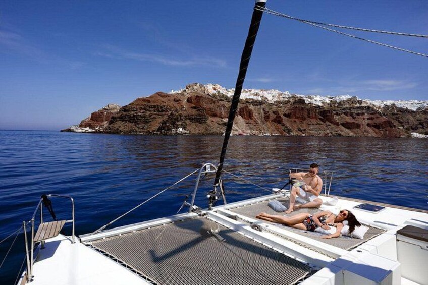 Eine Segelyacht mit zwei Personen, die entspannt auf dem Deck sitzen, eine felsige Küstenlinie und ruhiges, blaues Wasser. 2