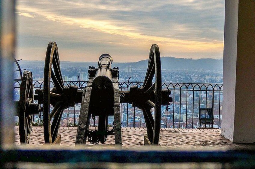A cannon with two large wheels, overlooking a cityscape with buildings and hills in the distance. 1