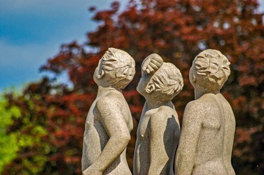 Tres estatuas de piedra de niños con hojas en la cabeza, frente a un fondo de árboles con hojas rojas. 1