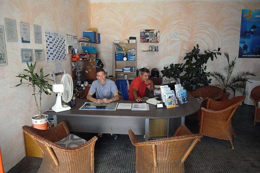 A reception area with wicker chairs, a desk, a fan, and various informational posters. 1