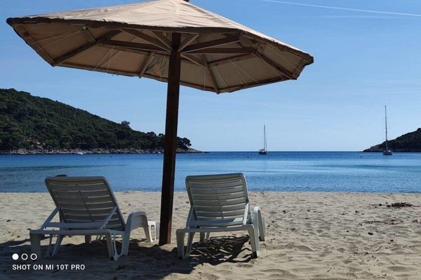 A beach with two lounge chairs under a large umbrella, a sandy shore, and sailboats in the distance. 2