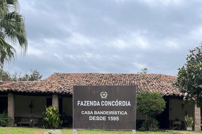 A building with a tiled roof, a sign reading 'FAZENDA CONCORDIA CASA BANEIRISTICA DESDE 1595,' and some greenery around. 3