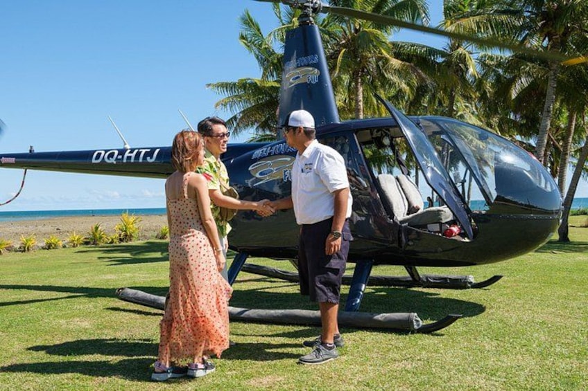A helicopter with 'DC-HTJ' on its tail, a man in a white shirt and cap, and a woman in a polka dot dress standing on grass near palm trees. 3