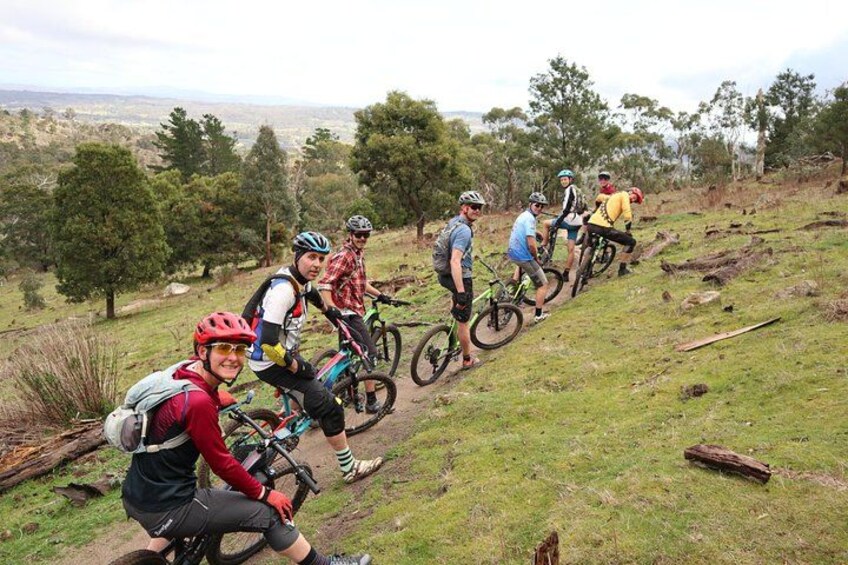A group of cyclists on a dirt trail with trees and a hilly landscape in the background. 1