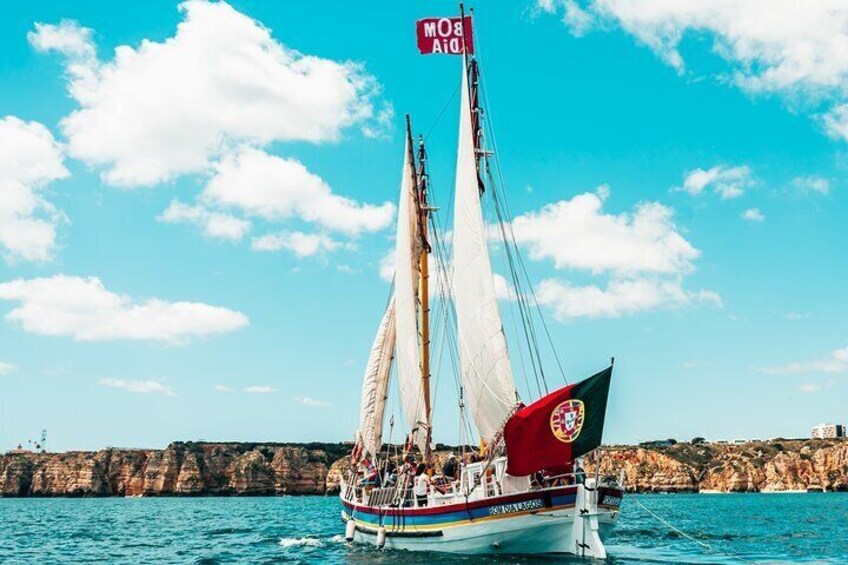 A sailboat with a red flag and a logo on the stern, sailing on turquoise water with a rocky coastline in the background. 2