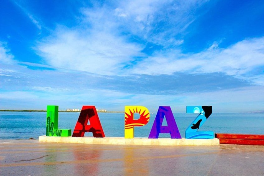 Colorful letters spelling 'LA PAZ' on a beach with a calm sea and clear sky in the background. 1