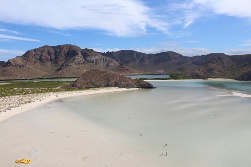A beach with clear water, a rocky outcrop, and mountains in the background. 3