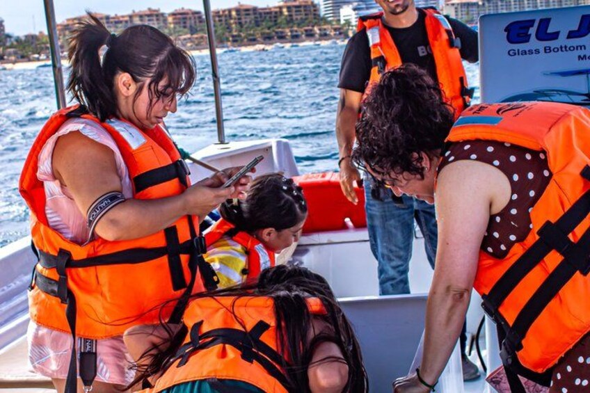 Personas en un bote con chalecos salvavidas anaranjados, un letrero que dice 'ELJ Glass Bottom Marina' y un cuerpo de agua con edificios al fondo. 5