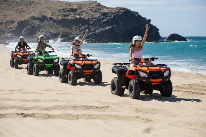 Three people riding ATVs on a sandy beach with rocky cliffs in the background. 1