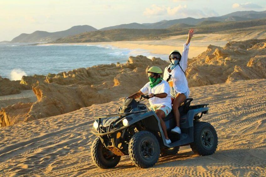Two people on an ATV in a desert with a beach and mountains in the background. 3