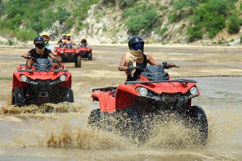 People riding red ATVs on a muddy riverbank. 4