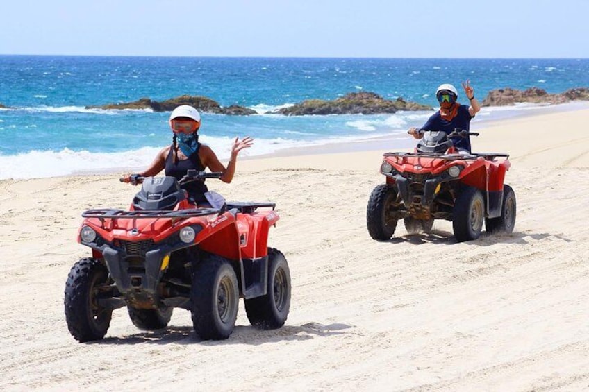 Two people riding red ATVs on a sandy beach with the ocean in the background. 2